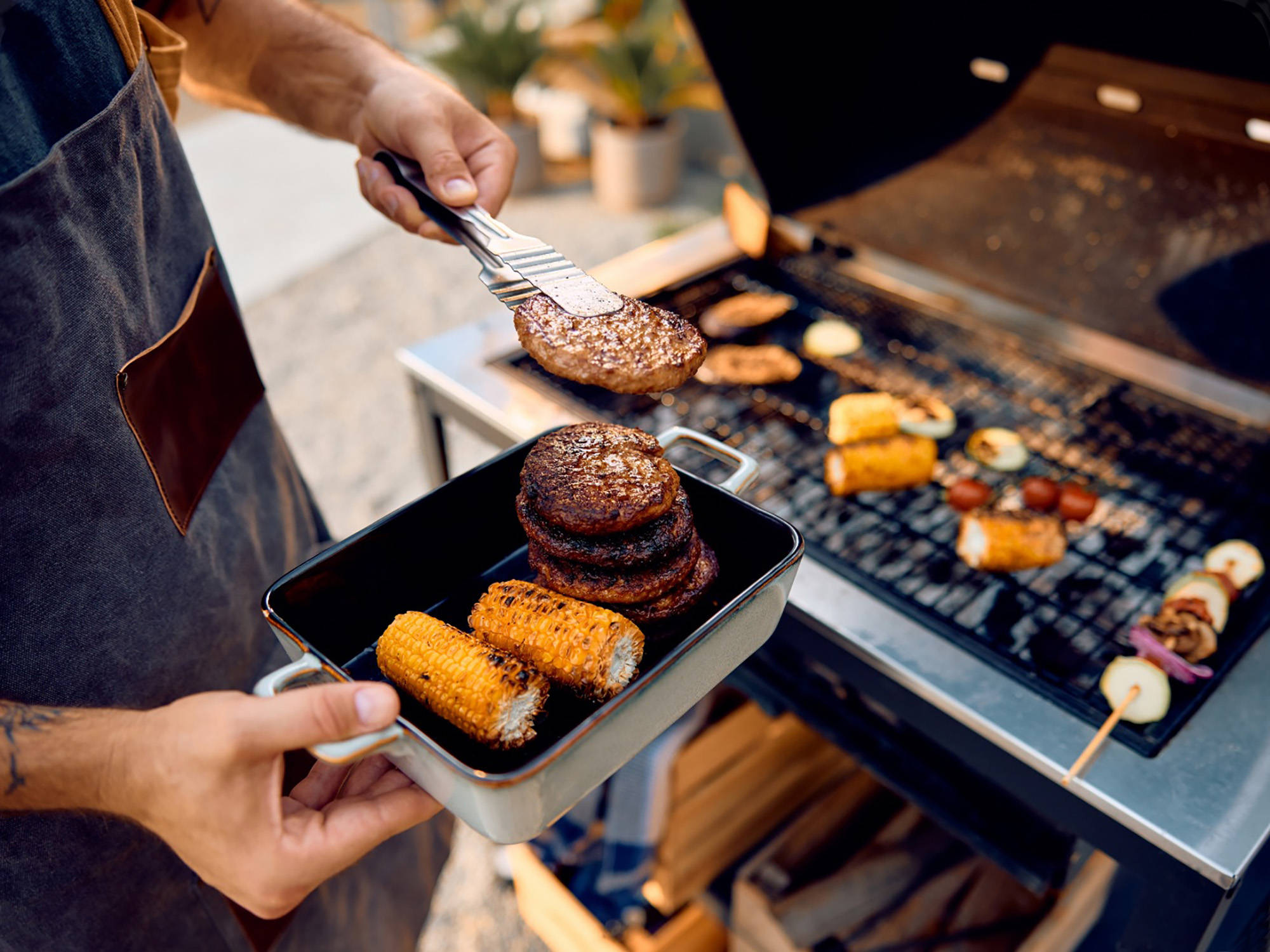 Person nimmt Grillgut aus Burger-Patties und Maiskolben vom Grill in Metallschale, daneben Gemüse auf Grillrost.