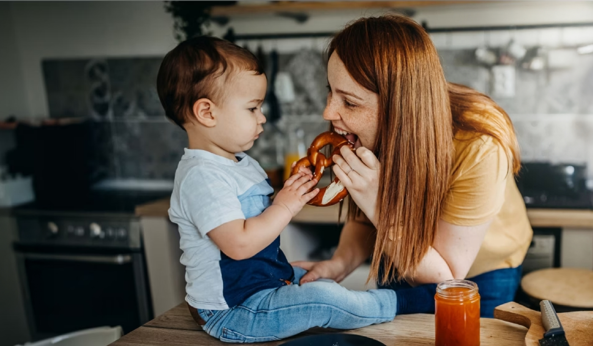 Kleinkind sitzt auf der Küchenarbeitsfläche und hält eine Brezel in den Händen, während die Mutter lachend hineinbeißt.
