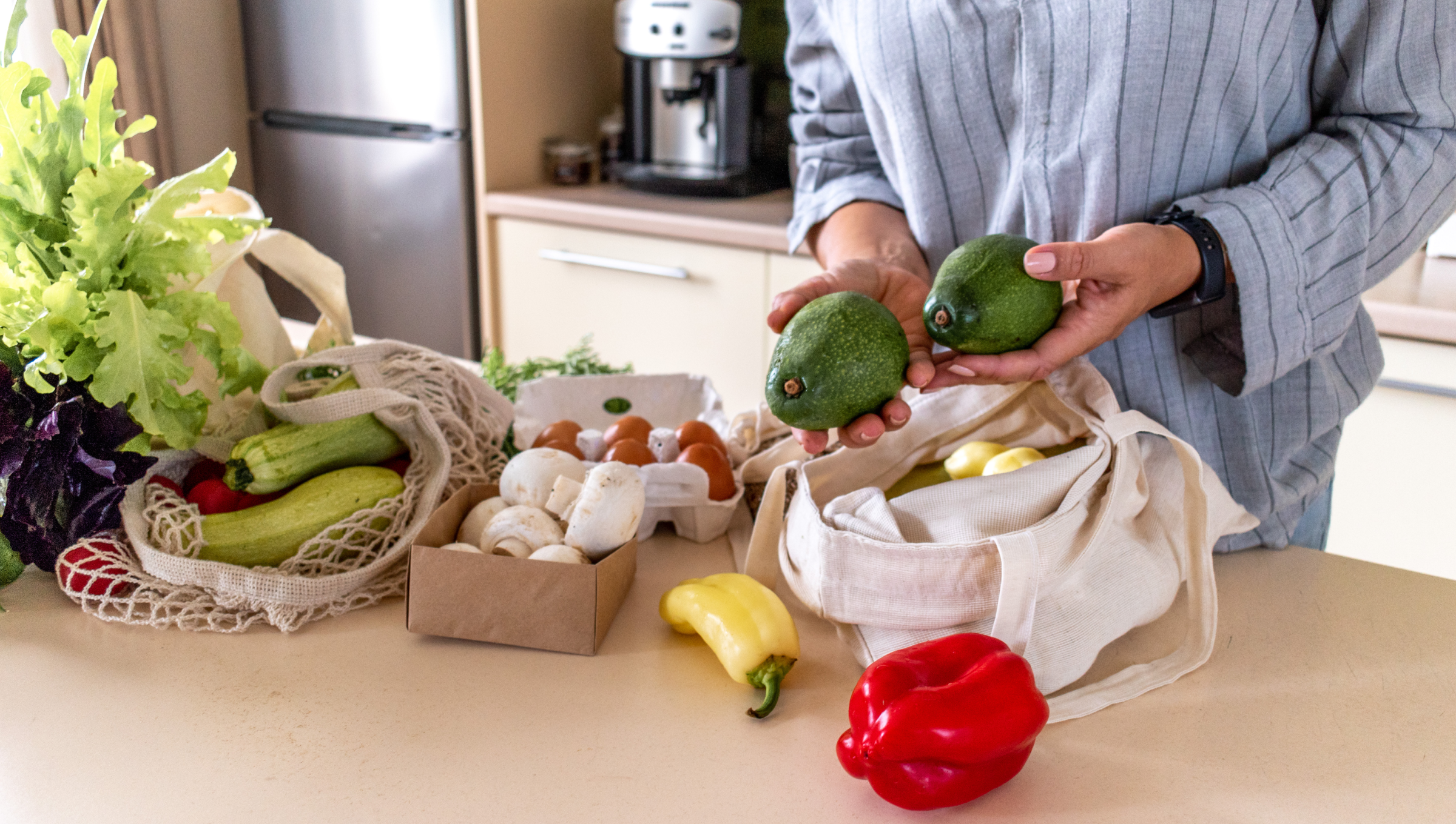 Person hält zwei Avocados über einem Tisch mit frischem Gemüse und Eiern in einer Küche.