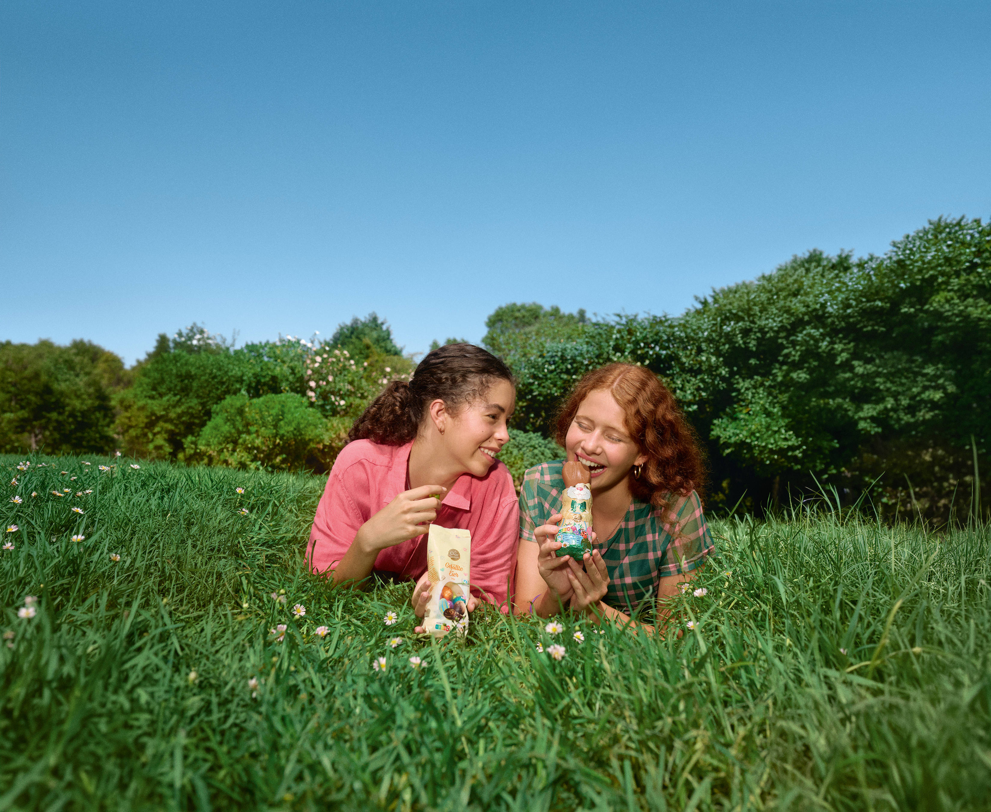 Zwei Mädchen liegen lachend auf einer grünen Wiese und essen Schoko-Osterhasen.