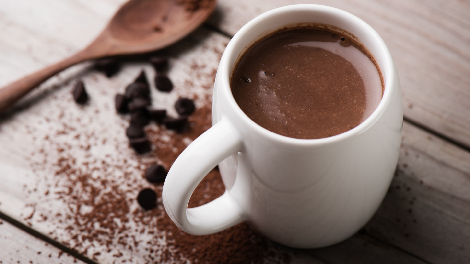 Schokomilch in weißer Tasse mit Schokostückchen und Holzlöffel.