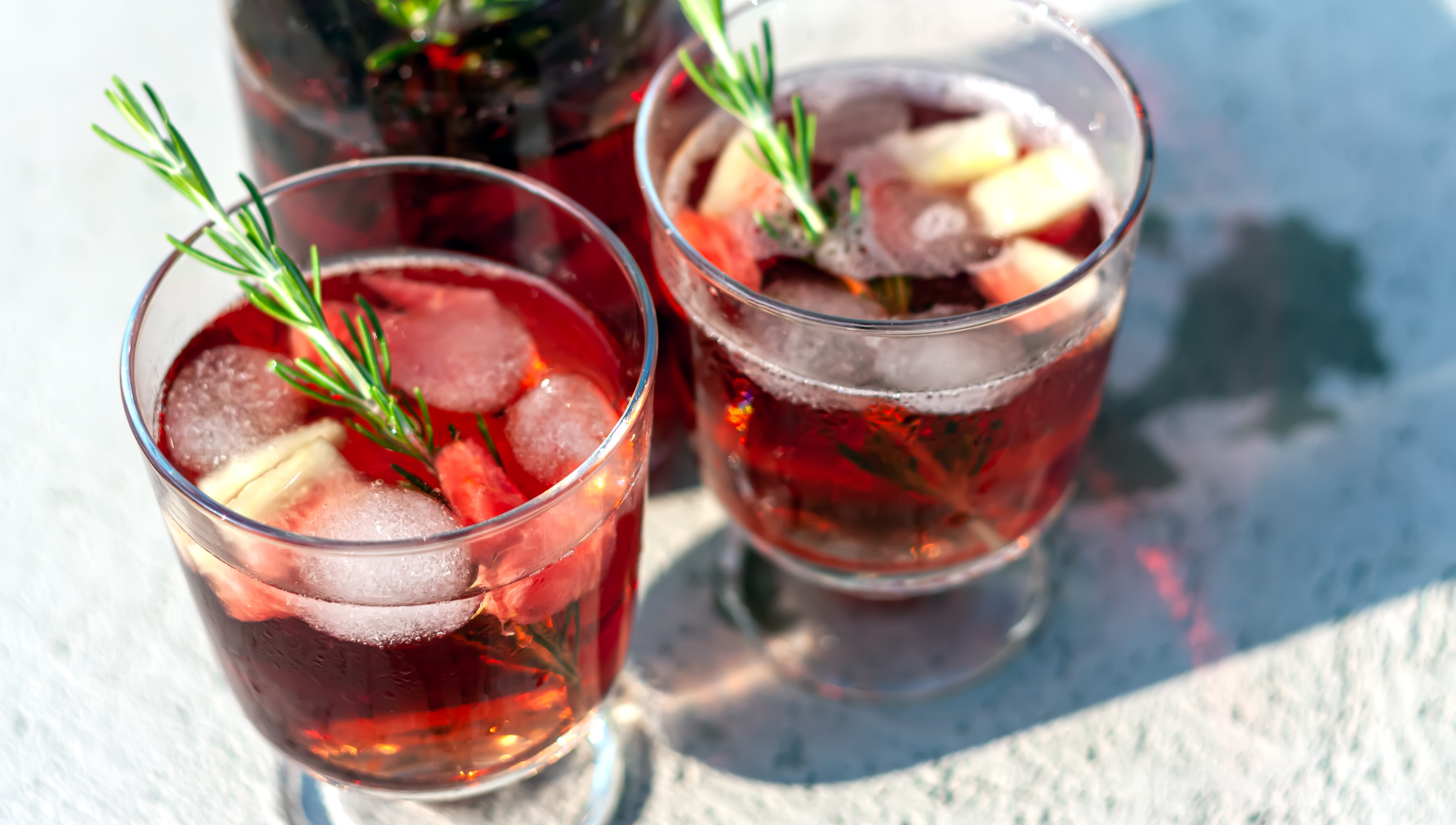 Zwei Gläser roter Sommerdrink mit Eiswürfeln, Rosmarin, Erdbeeren und Ingwer auf einem Tisch in der Sonne.
