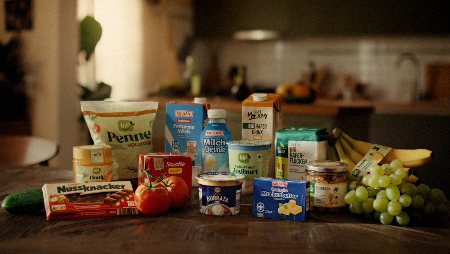 Verschiedene Lebensmittel von ALDI SÜD wie Pasta, Milch, Haferdrink, Butter, Obst und Gemüse auf einem Küchentisch.