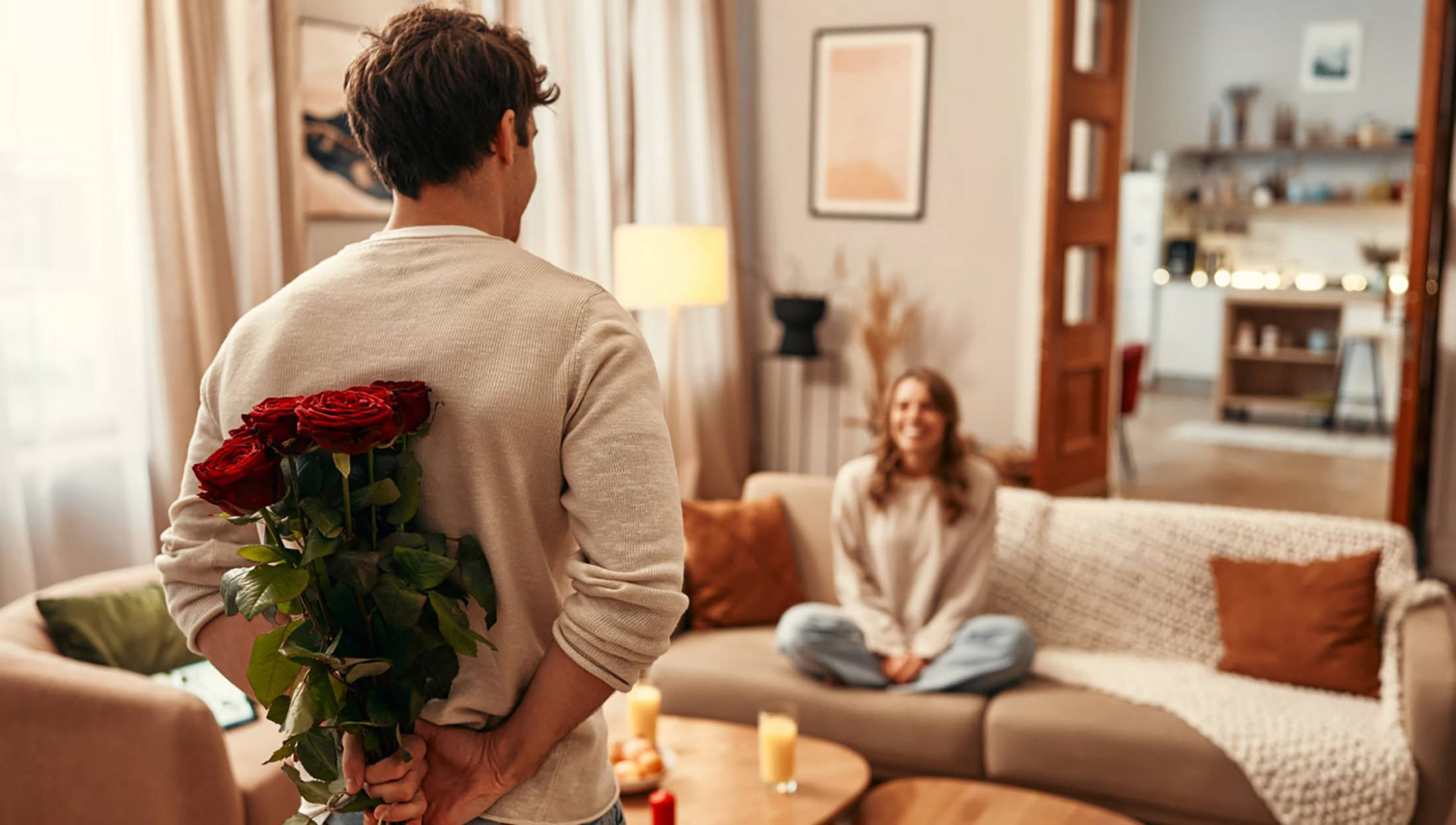 Mann versteckt einen Rosenstrauß hinter dem Rücken, während eine Frau lächelnd auf einem Sofa in einem gemütlichen sitzt