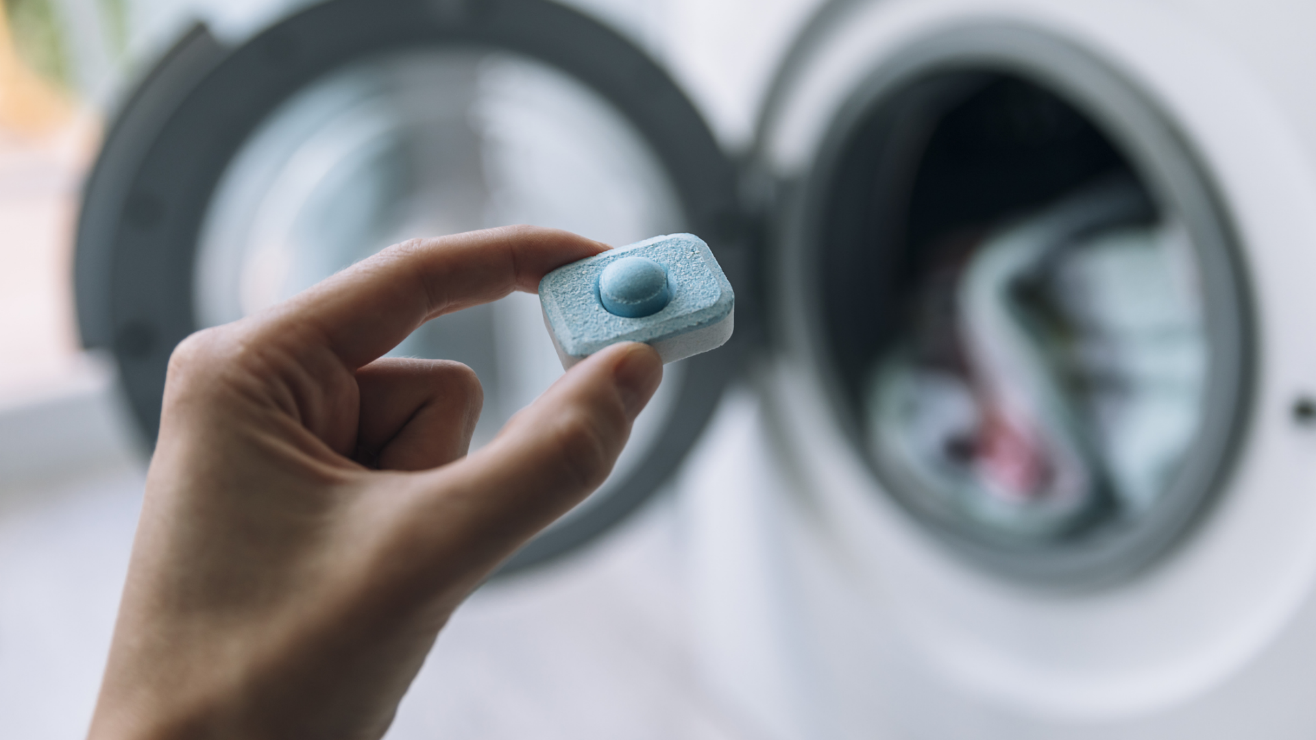 Hand hält Waschmittel-Tab vor Waschmaschine.