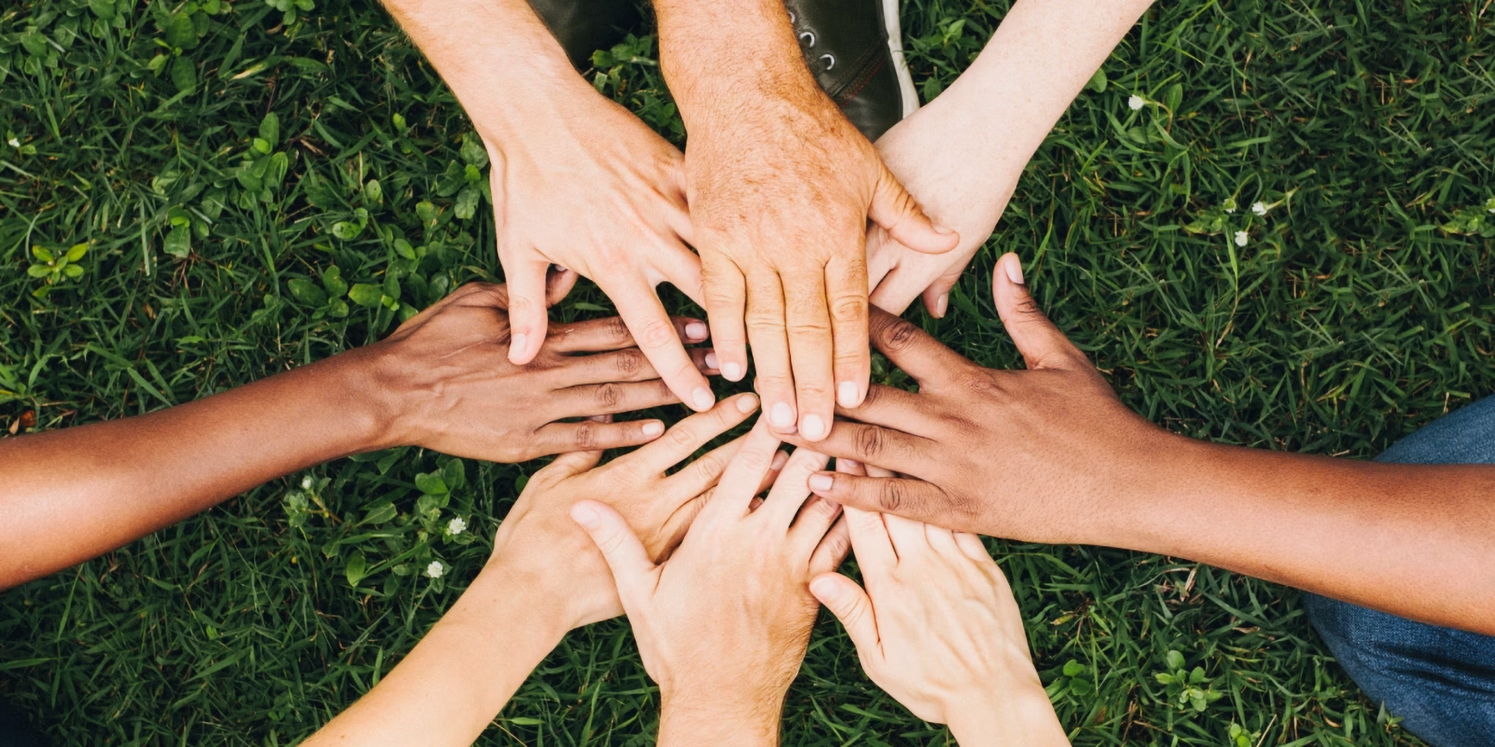 Verschiedene Hände legen sich aufeinander im Kreis auf grünem Gras.