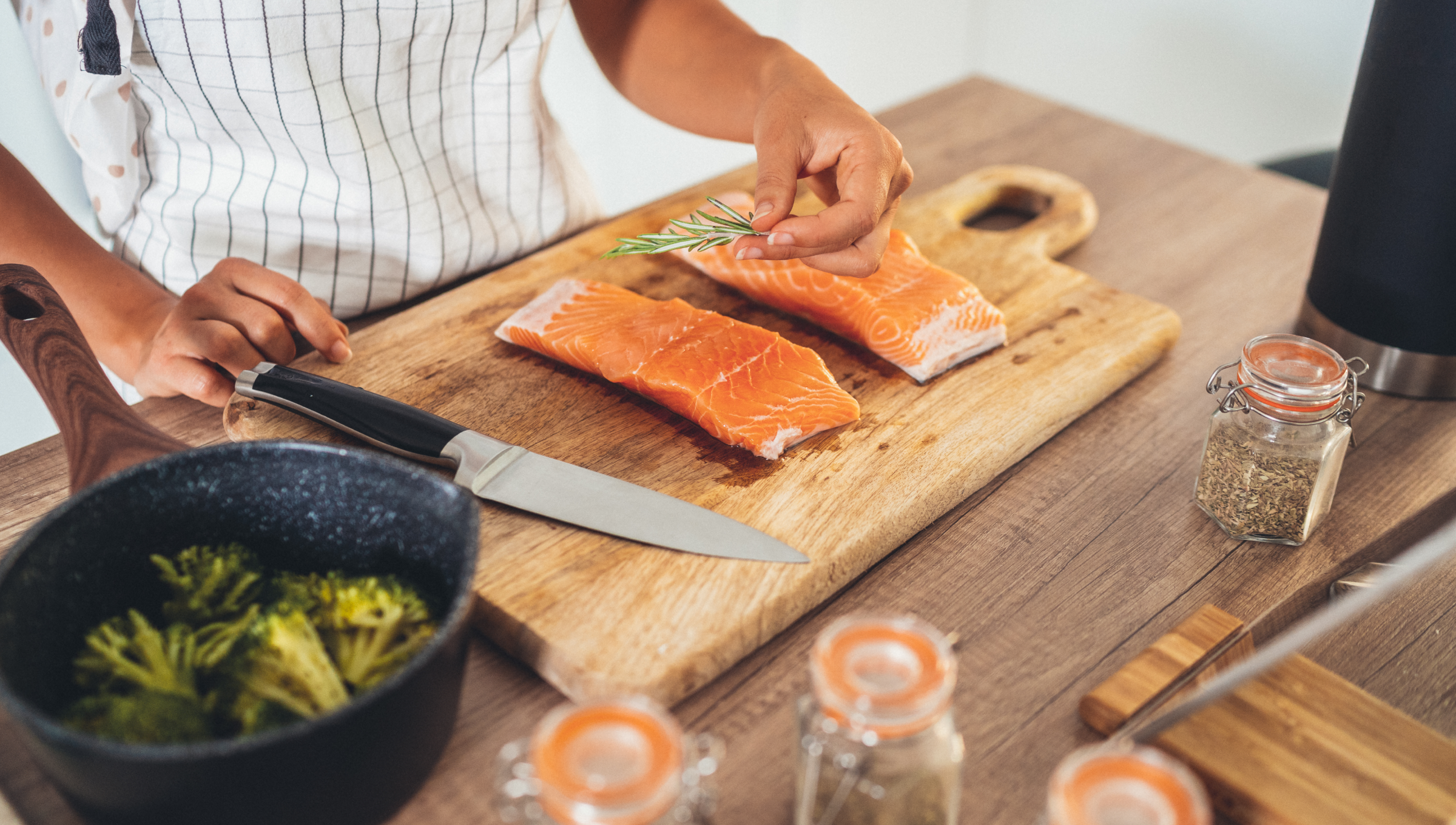 Eine Person bereitet zwei Lachsfilets auf einem Küchenbrett zu. Daneben frische Gewürze und ein Kochtopf.