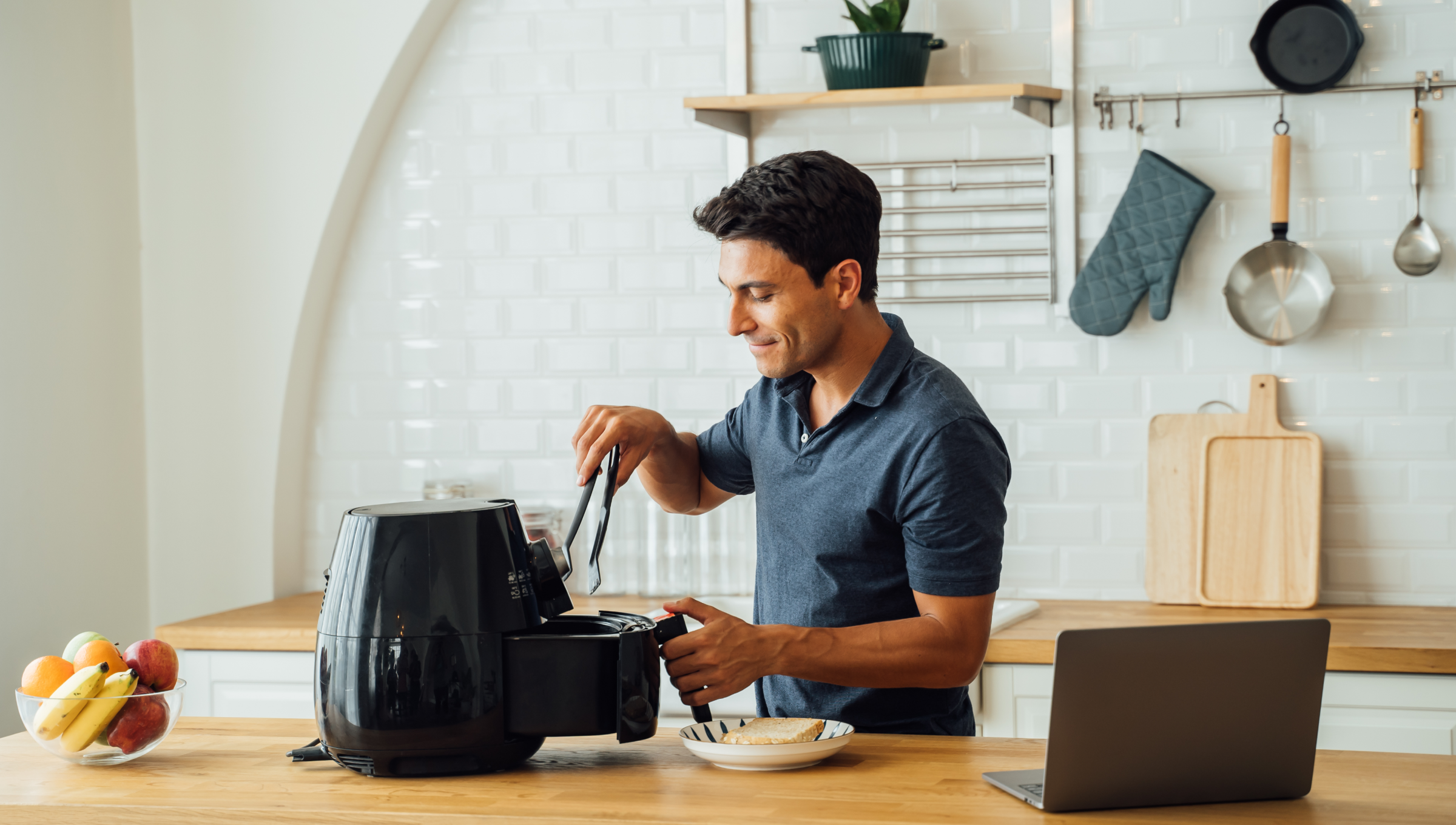 Ein Mann steht mit einer Küchenzange in der Hand vor einer geöffneten Heißluftfritteuse und schaut hinein.