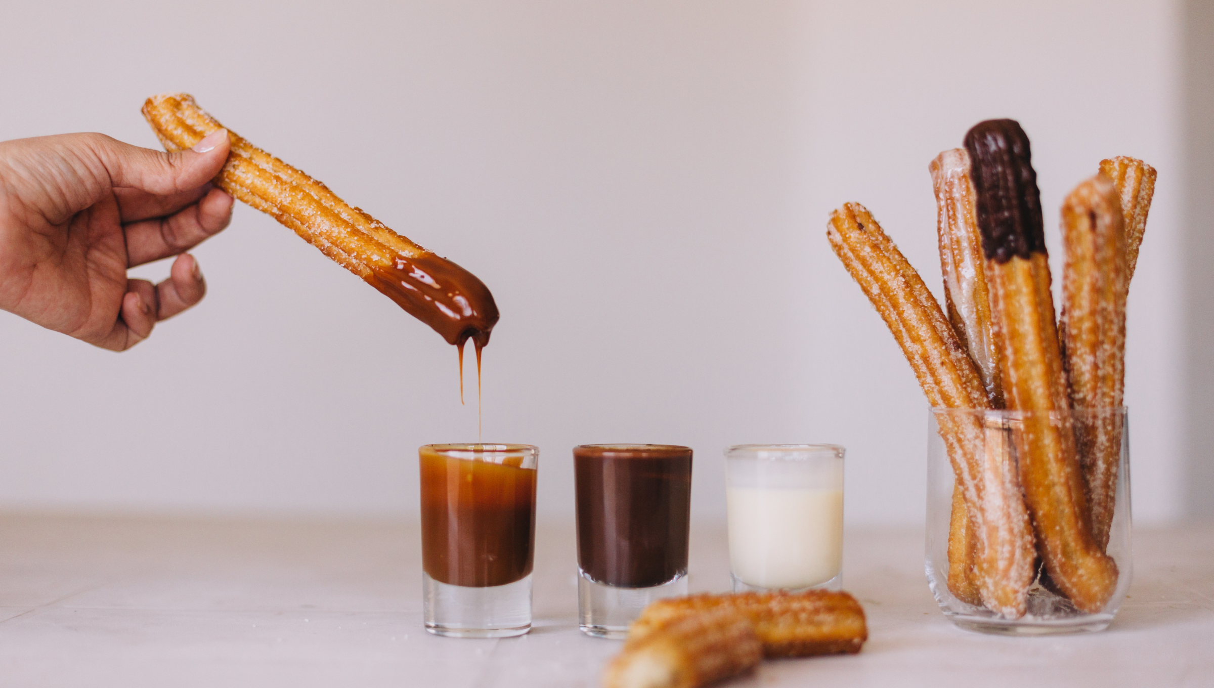 Jemand taucht einen Churro in flüssige Schokolade, daneben stehen Gläser mit Karamell und Sahne und weitere Churros.