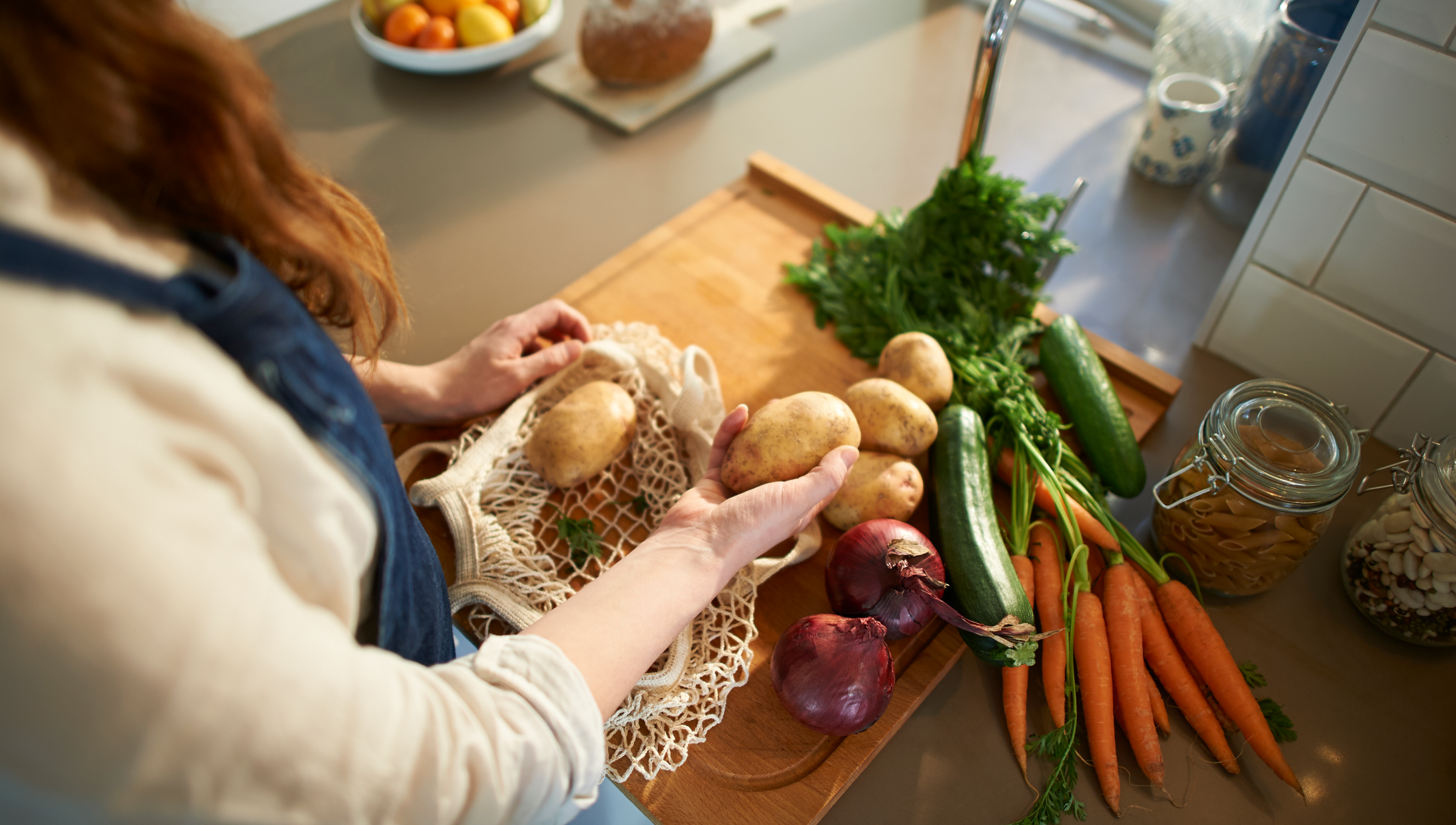 Eine Frau legt Kartoffeln aus einem Netzbeutel auf eine Küchenarbeitsplatte mit frischem Gemüse.