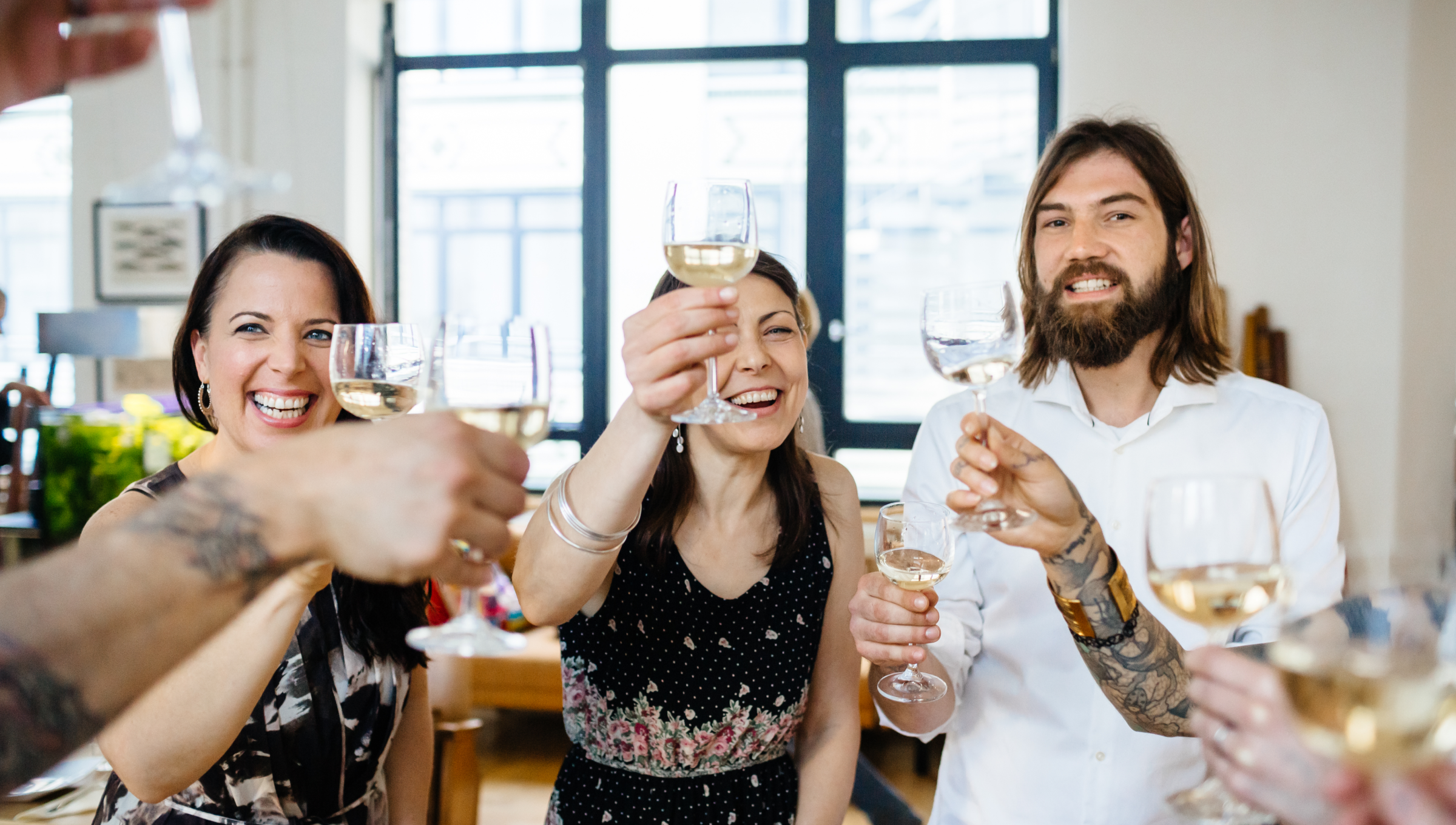 Gruppe von Freunden stößt mit Weißweingläsern in einem hellen Raum an, alle lächeln und sind fröhlich.